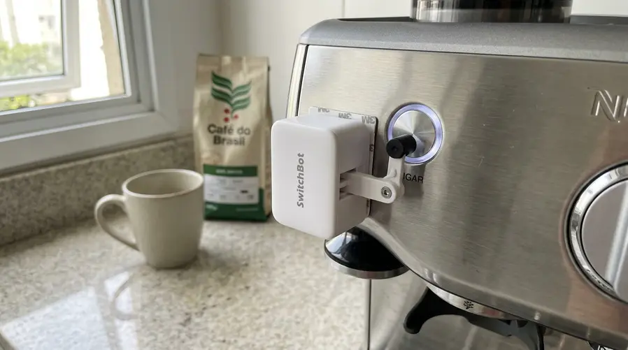 A imagem deve mostrar o Bot Rechargeable em uso real ao lado de uma cafeteira ou de um botão físico de aparelho doméstico, com foco no acessório pressionando um botão comum, de preferência dentro de uma casa organizada, para reforçar a ideia de automação simples e útil no dia a dia.