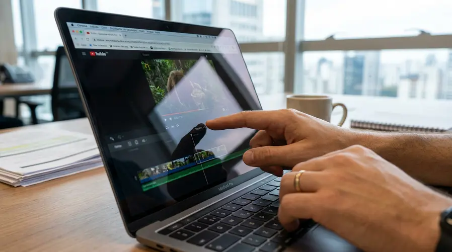 Um close de uma mão tocando a tela de um notebook Apple em uma cena de uso real, mostrando uma interface com vídeo em reprodução e uma linha do tempo de edição ou uma página aberta em navegador, para ilustrar tarefas do dia a dia que ganhariam agilidade com o toque.