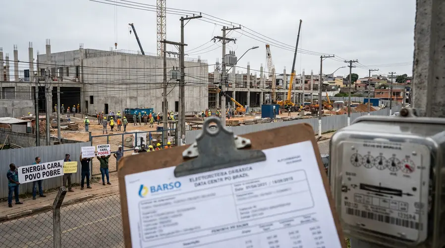Uma imagem de um grande data center em construção ao lado de postes e linhas de energia, com placas de protesto ao fundo e um medidor de energia ou fatura de luz em primeiro plano, para ilustrar visualmente a pressão sobre a conta de energia e a infraestrutura.