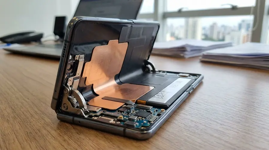 Uma imagem ou ilustração do interior de um smartphone dobrável mostrando camadas de dissipação de calor, com destaque para a região da placa e da bateria, além de uma dobra aberta ao lado sugerindo a dobradiça reforçada. A ideia é visualizar o mecanismo de controle térmico e não apenas o aparelho por fora.