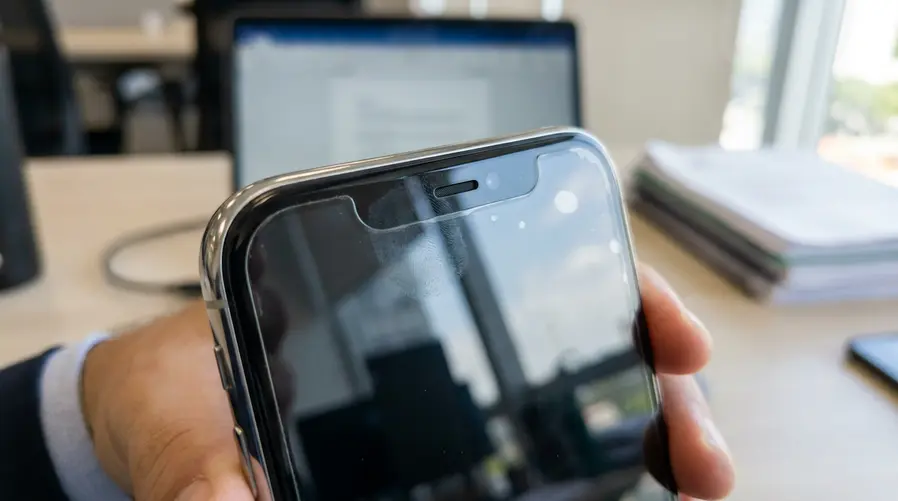 Uma imagem aproximada da parte superior do iPhone mostrando a área da câmera TrueDepth com uma película mal alinhada ou com marcas de sujeira/oleosidade na borda da tela, para evidenciar visualmente a obstrução que pode atrapalhar o Face ID.