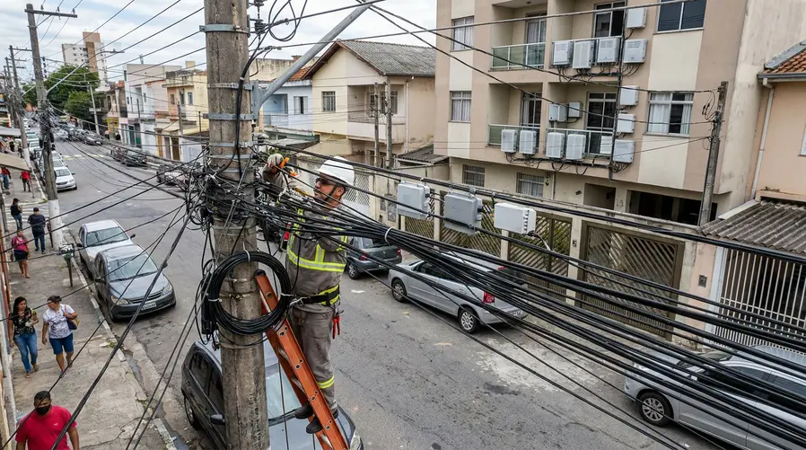 Uma cena de rua urbana brasileira com vários cabos e postes cheios de fiação, enquanto um técnico sobe em uma escada para organizar cabos de fibra óptica; ao fundo, casas e um prédio residencial com caixas de internet na fachada, ilustrando a disputa pelo uso dos postes e o efeito disso na chegada da fibra ao consumidor.