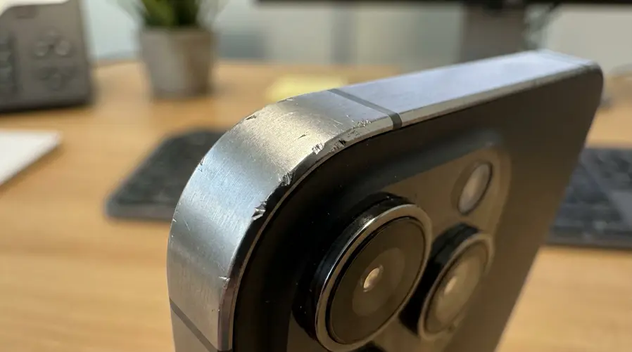 Um close realista da borda superior da ilha da câmera de um iPhone Pro com pequenas marcas, riscos e lascas na quina do alumínio anodizado, destacando visualmente o ponto mais vulnerável sem mostrar o aparelho inteiro.