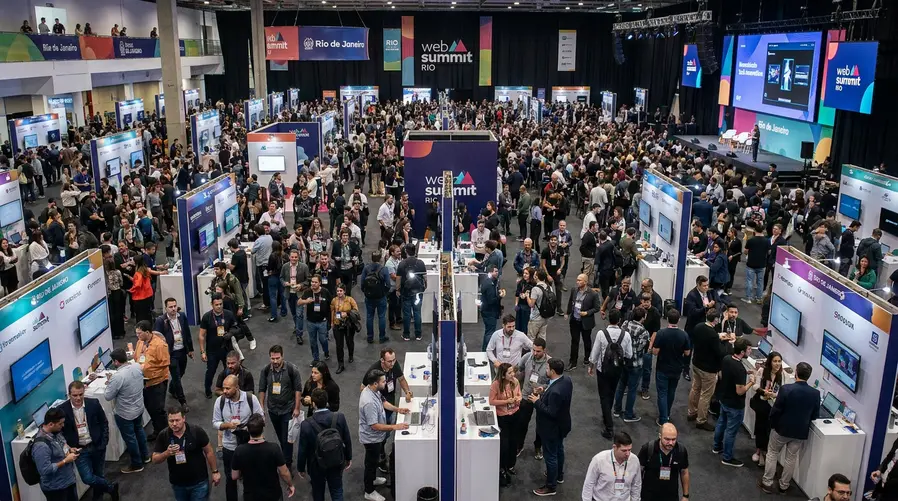 Uma foto ampla do auditório ou área principal do Web Summit Rio com grande fluxo de pessoas, estandes de startups e placas do evento ao fundo, transmitindo a escala citada pela seção. Se possível, incluir elementos visuais de tecnologia e networking, como pessoas conversando em frente a telas e expositores.